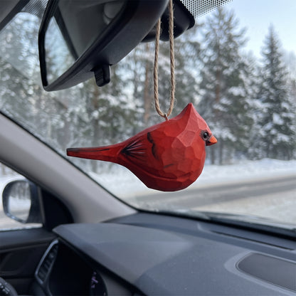 A red geometric bird-shaped car air freshener hanging from a car's rearview mirror, with a snowy forest road visible through the windshield.