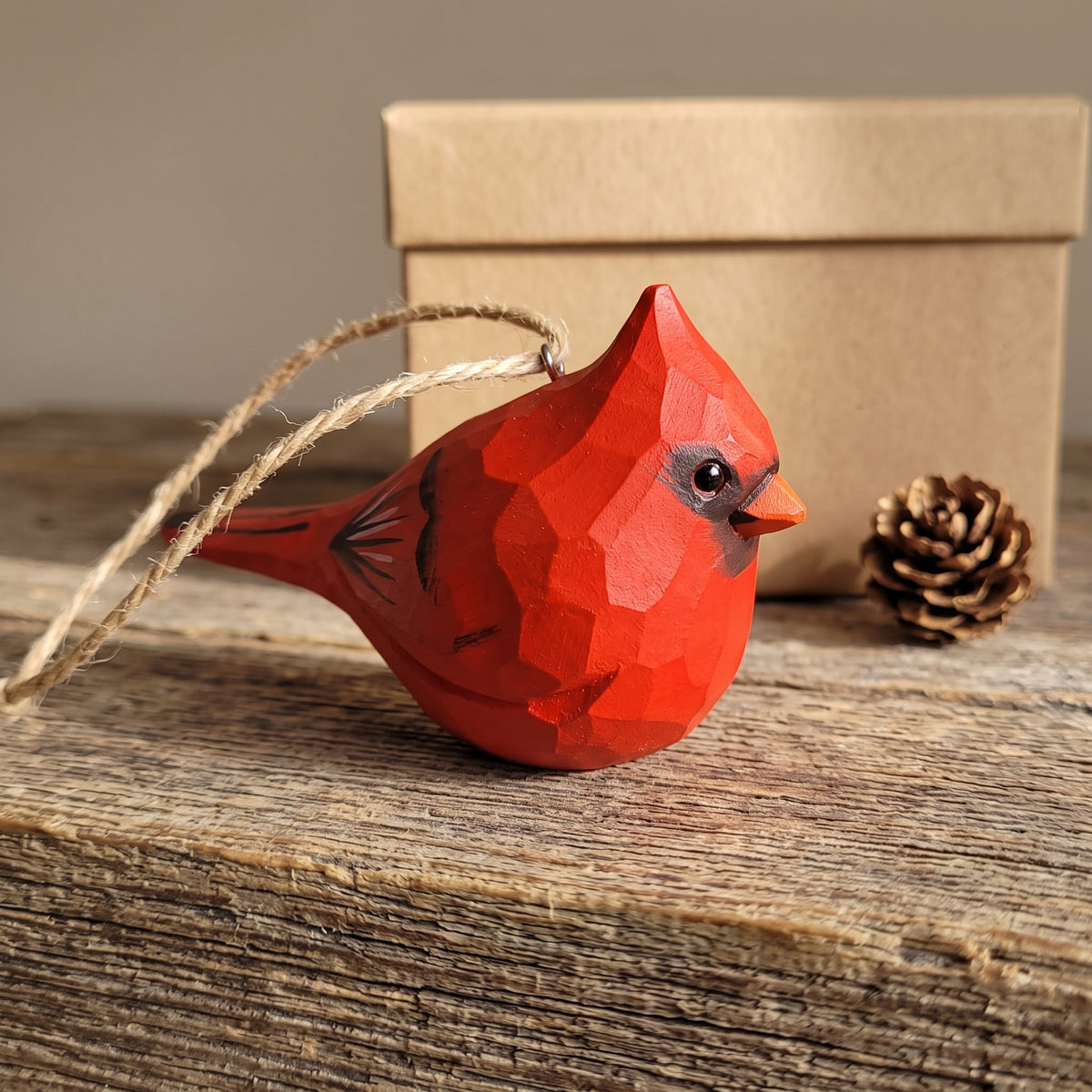 A red geometric bird ornament with a hanging twine loop, displayed on a rustic wooden surface with a small pinecone and a kraft gift box in the background.