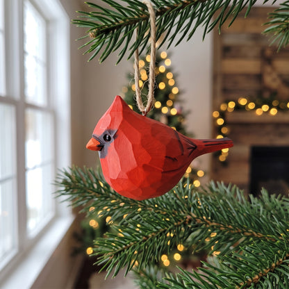 A red geometric cardinal bird Christmas ornament hanging from a pine tree branch in a cozy indoor setting with blurred warm lights and a window in the background.