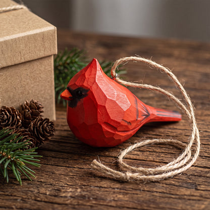 A red geometric cardinal Christmas ornament with a natural jute hanging string, displayed on a rustic wooden surface next to pinecones and a kraft gift box.