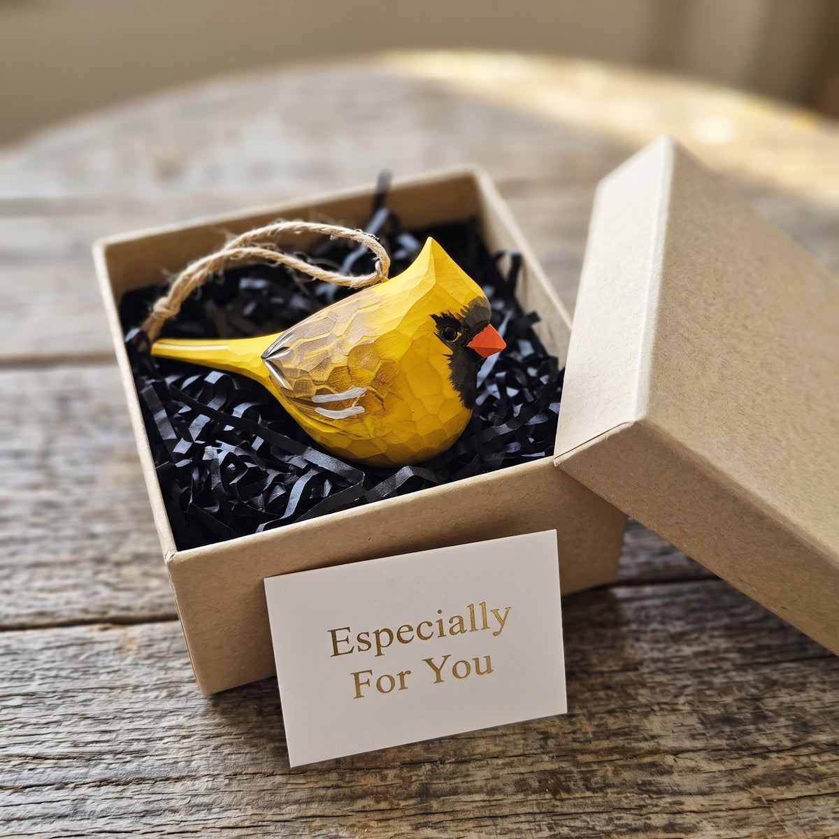A yellow bird-shaped ornament with a textured geometric pattern sits in a brown kraft gift box filled with black shredded paper. A small card reading 'Especially For You' is placed in front, and the box is set on a rustic wooden surface.