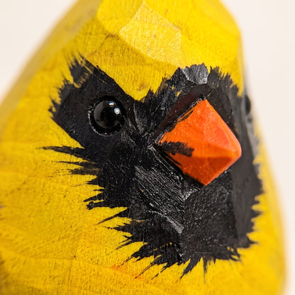 Close-up of a carved wooden Yellow Cardinal figurine, likely a cardinal, with a bright yellow body, black painted feathers, and a small orange beak on a plain light background.