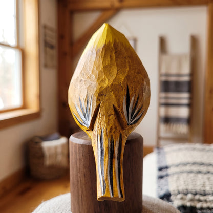 A hand-carved wooden Yellow Cardinal sculpture of a stylized animal head, likely a bison or antelope, in warm natural wood tones with black and white painted details, displayed on a wooden pedestal in a rustic indoor setting.