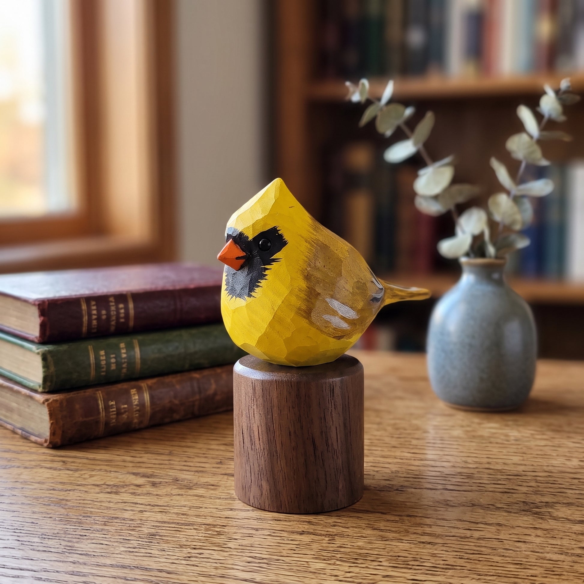A hand-carved wooden Yellow Cardinal figurine in bright yellow with black, orange, and white facial details, mounted on a round dark wood base on a wooden table. The background features a blurred bookshelf, a stack of vintage books, and a small vase with dried eucalyptus branches.