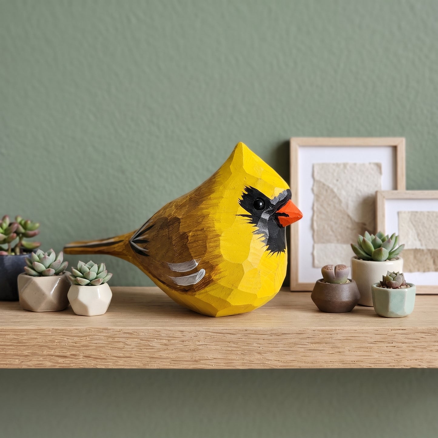 A geometric, faceted wooden Yellow Cardinal sculpture in bright yellow, brown, black, and orange, displayed on a light wood shelf alongside small succulent plants and framed art against a sage green wall.