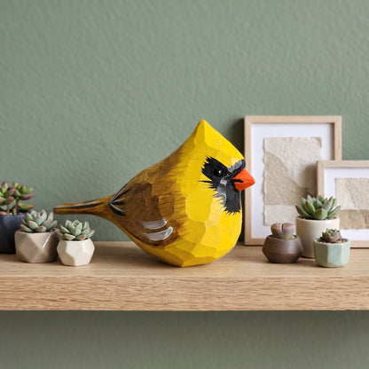 A geometric, faceted wooden Yellow Cardinal sculpture in bright yellow, brown, black, and orange, displayed on a light wood shelf alongside small succulent plants and framed art against a sage green wall.