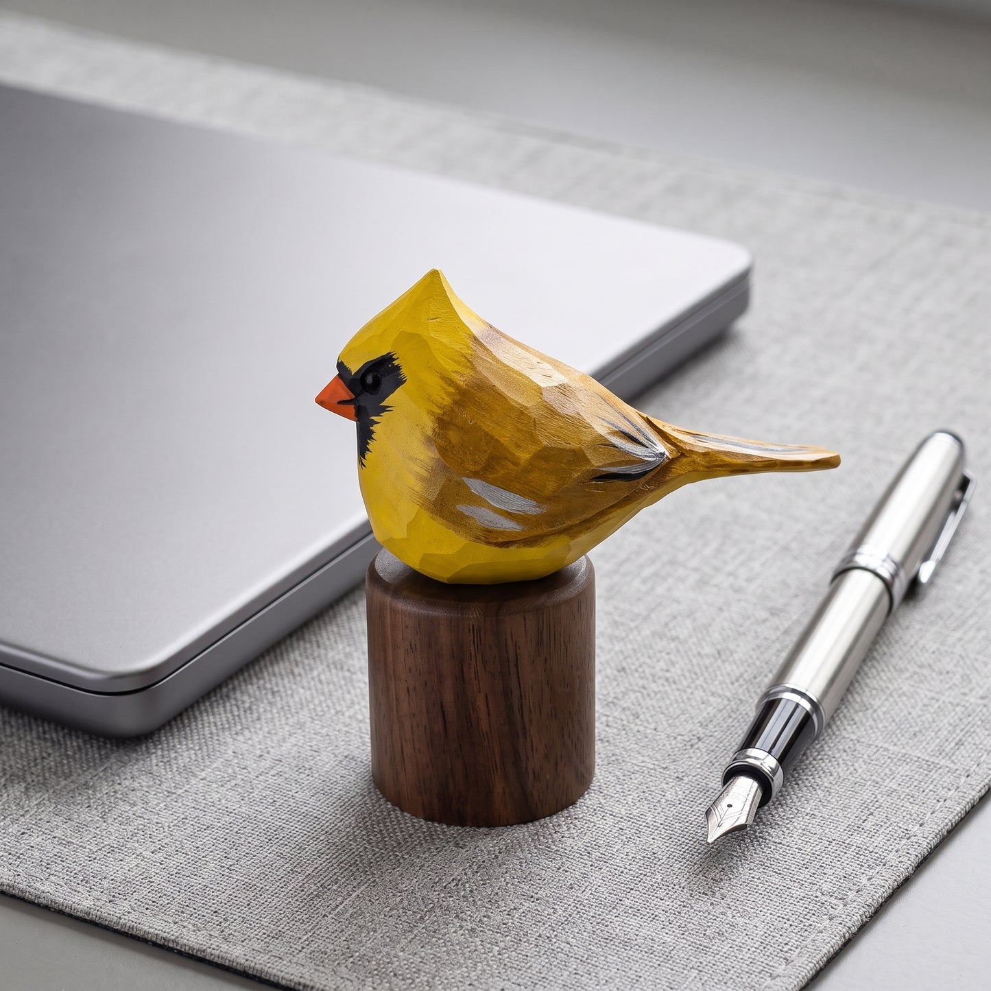 A carved wooden Yellow Cardinal figurine painted in yellow, black, and orange, mounted on a dark wood cylinder base. It is styled on a gray textured desk mat next to a silver laptop and a silver fountain pen.