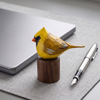 A carved wooden Yellow Cardinal figurine painted in yellow, black, and orange, mounted on a dark wood cylinder base. It is styled on a gray textured desk mat next to a silver laptop and a silver fountain pen.