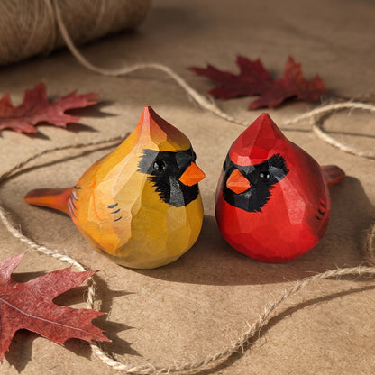 Two hand-painted wooden cardinal figurines, one yellow and one red, displayed on a rustic brown paper background with twine and autumn leaves.
