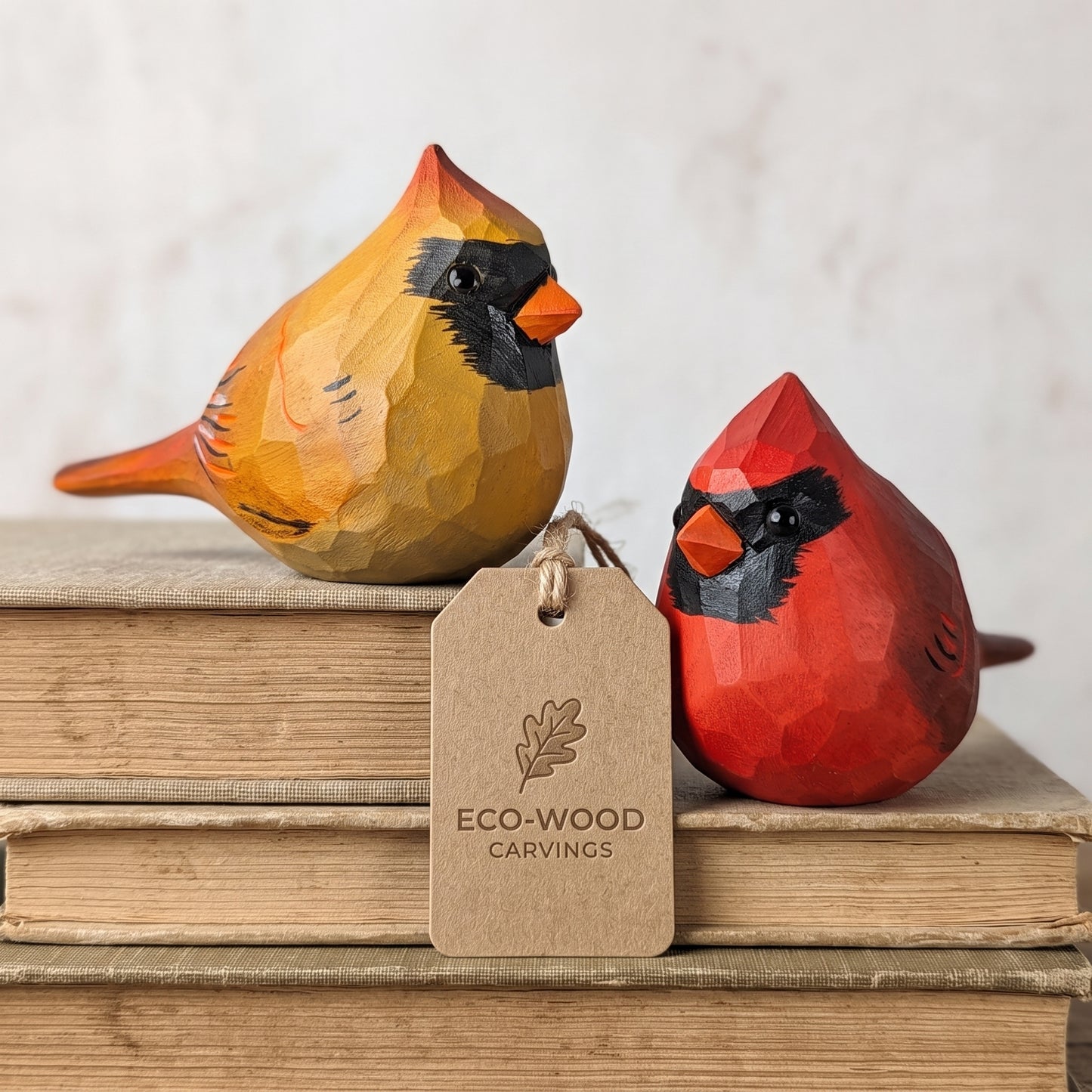 Two hand-carved wooden cardinal figurines, one yellow-orange and one red, displayed on a stack of vintage books with a hanging kraft paper tag that reads 'ECO-WOOD CARVINGS'.