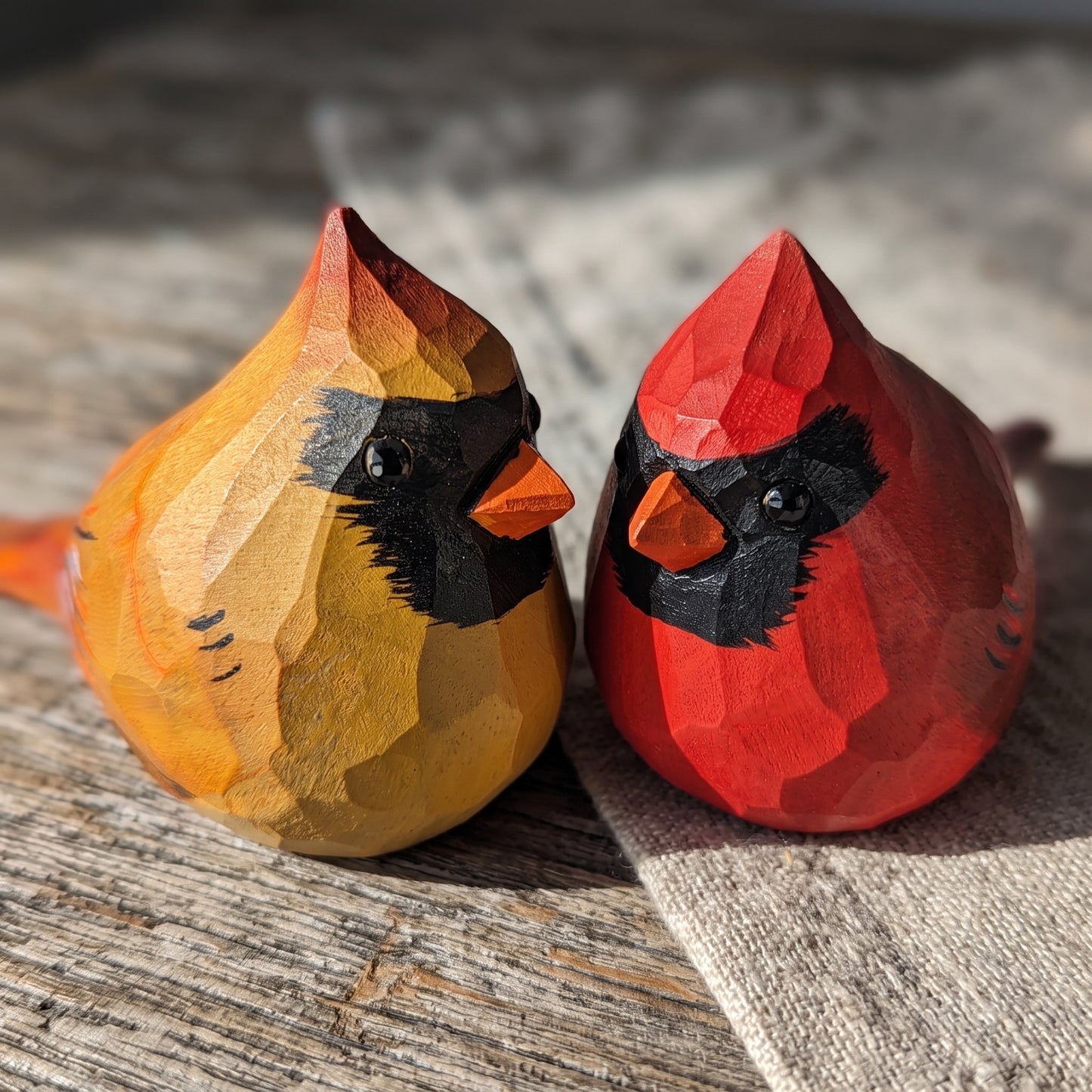 Two hand-carved wooden cardinal figurines, one yellow and one red, resting on a rustic wooden surface with a linen cloth.