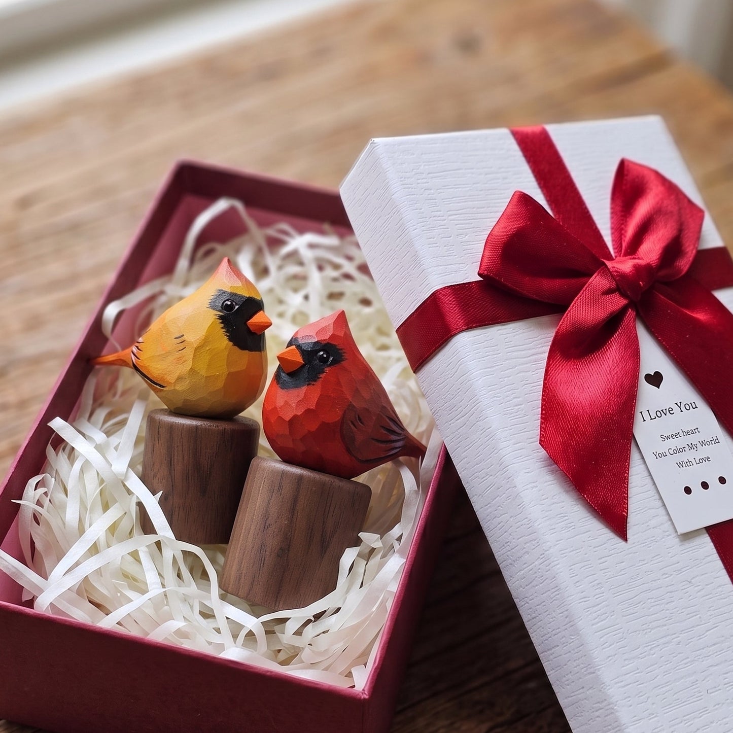 A gift box containing two wooden cardinal figurines, one orange-yellow and one red, displayed next to its white lid wrapped with a red ribbon on a rustic wooden table.