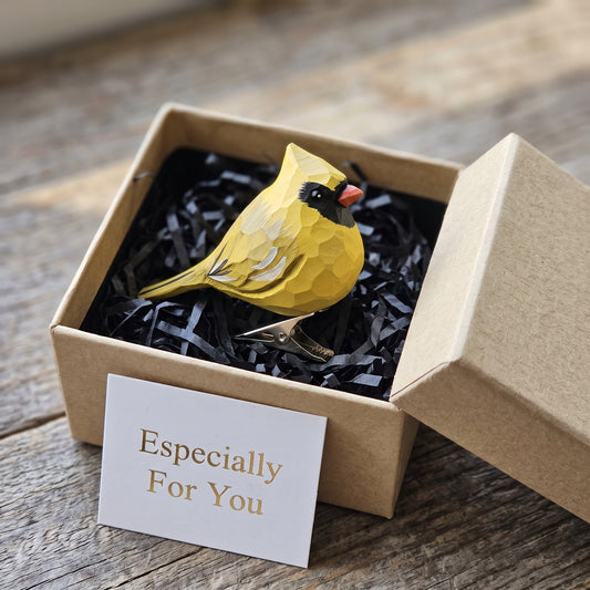 A hand-carved wooden Yellow Cardinal clip-on ornament displayed inside an open kraft gift box filled with black shredded paper, featuring a bright yellow body and crest, a bold black mask and bib, a silver metal clip attached underneath, and a white card reading "Especially For You" placed in the foreground.