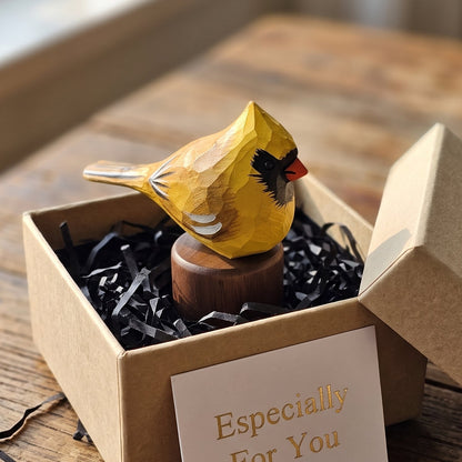 A handcrafted wooden Yellow Cardinal figurine painted bright yellow, perched on a small wooden base inside an open kraft gift box with black shredded paper and a card that reads 'Especially For You,' set on a rustic wooden table.