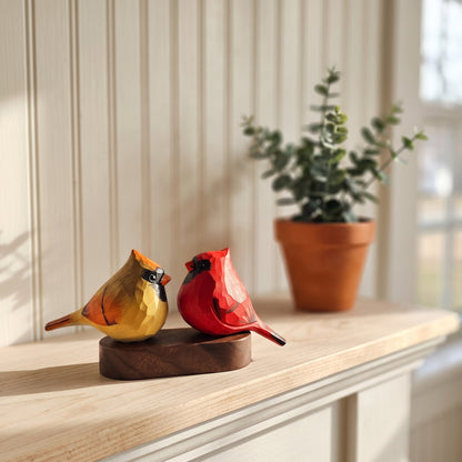 Two handcrafted wooden cardinal bird figurines, one yellow and one red, sitting side by side on a dark wooden base on a light wood mantelpiece, with a potted eucalyptus plant in the background.