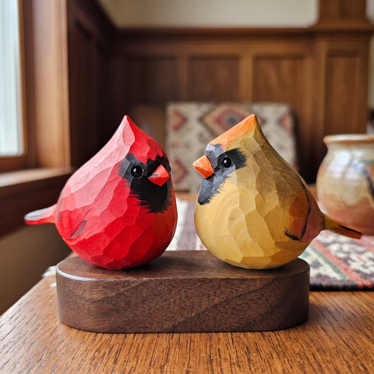 Two hand-carved wooden Cardinal Couple figurines on a round wooden base, set on a wooden table in a warm, wood-paneled room.
