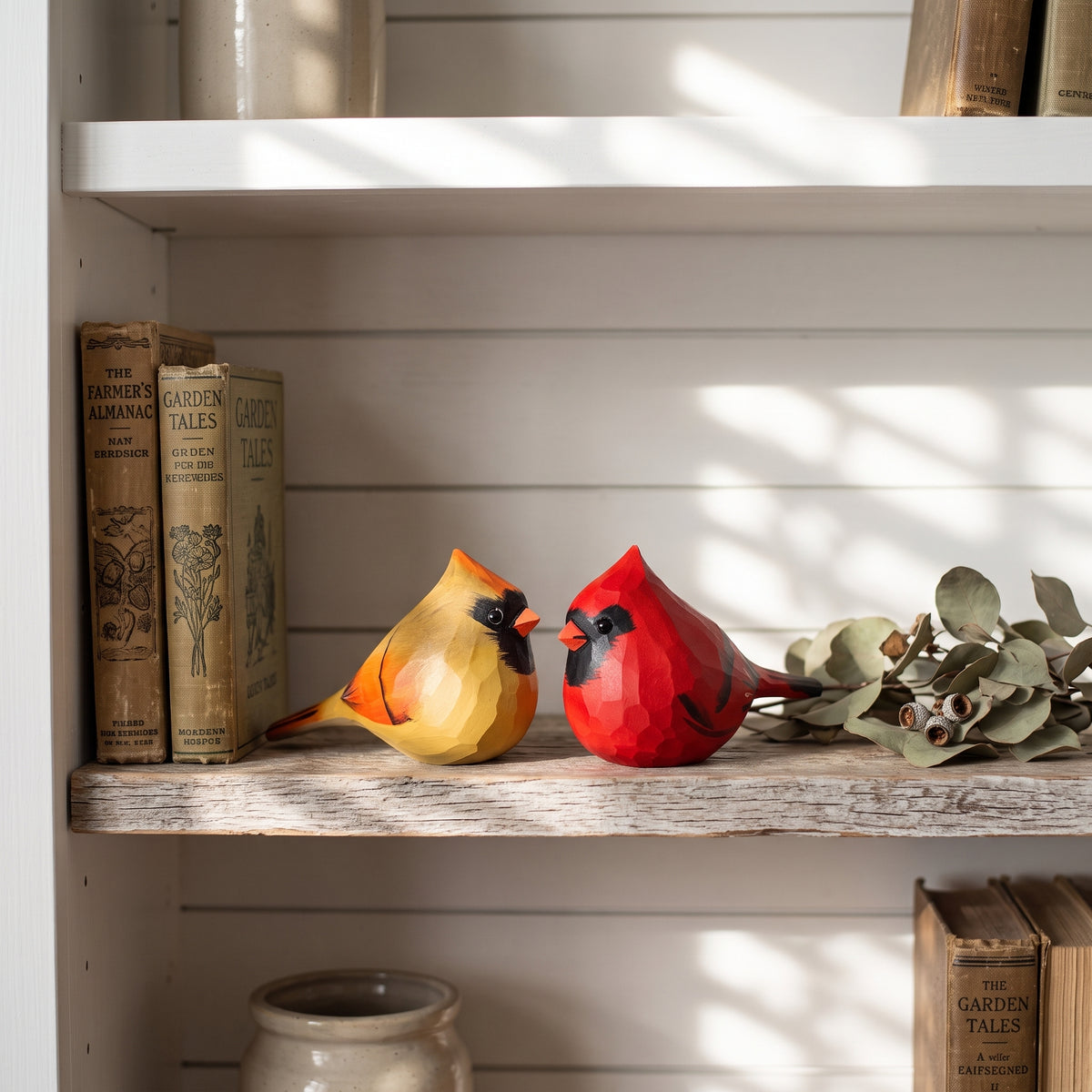 Two hand-painted wooden cardinal bird figurines, one yellow and one red, displayed on a rustic wooden shelf next to vintage books and eucalyptus branches.