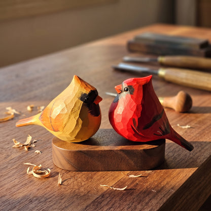 Two hand-carved wooden cardinal figurines, one yellow and one red, facing each other on a wooden base on a workbench with carving tools in the background.