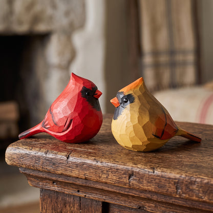 Two carved wooden cardinal bird figurines, one red and one yellow-brown, displayed on a rustic wooden mantel in a cozy indoor setting.