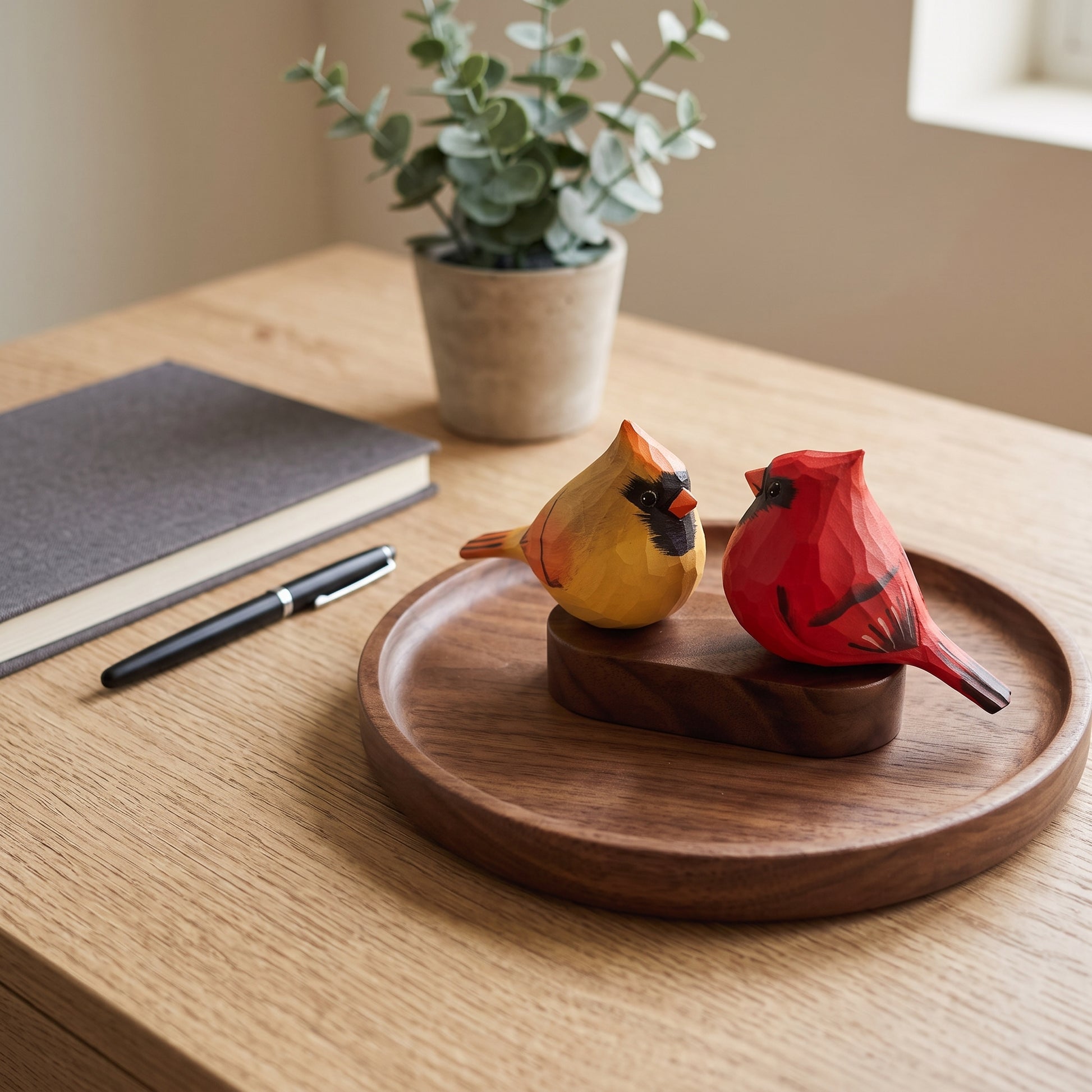 Two hand-carved wooden cardinal figurines, one yellow and one red, displayed on a round wooden tray on a light wood desk next to a notebook and pen.