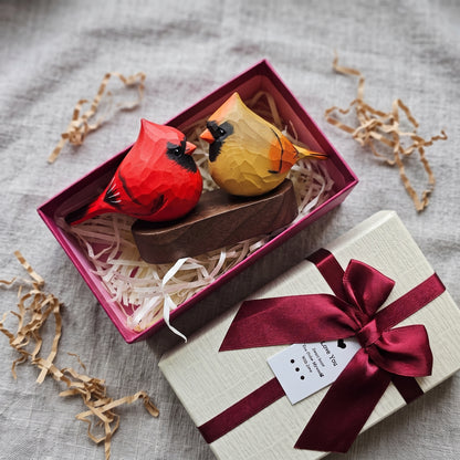 A gift set featuring two faceted origami cardinals, one red and one yellow, resting in a magenta gift box with shredded paper. A cream-colored gift box with a dark red satin ribbon and bow is shown next to it on a textured gray fabric background.