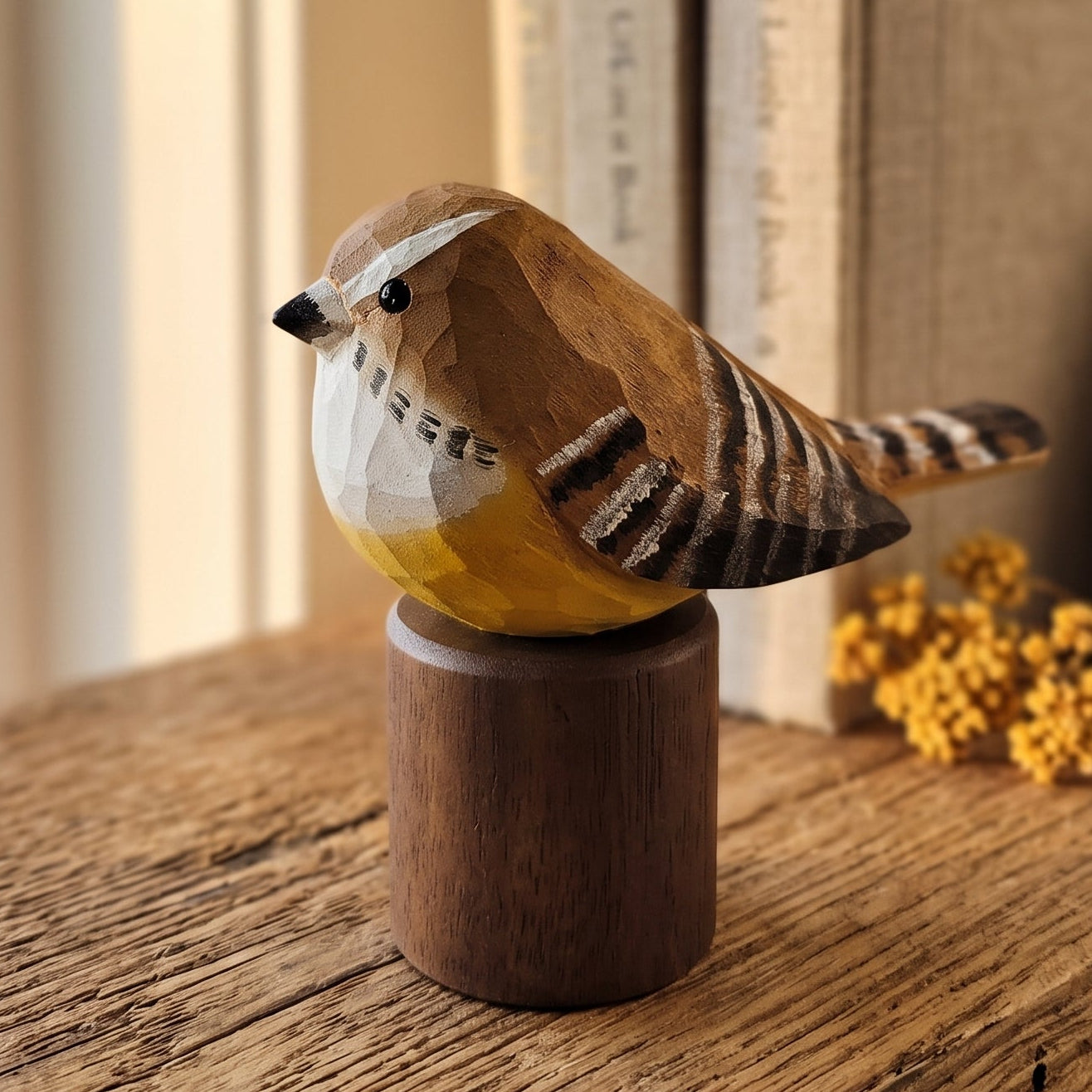 A handcrafted wooden Carolina Wren figurine with geometric facets sits on a cylindrical wooden base on a rustic wooden table, with books and a vase blurred in the background.