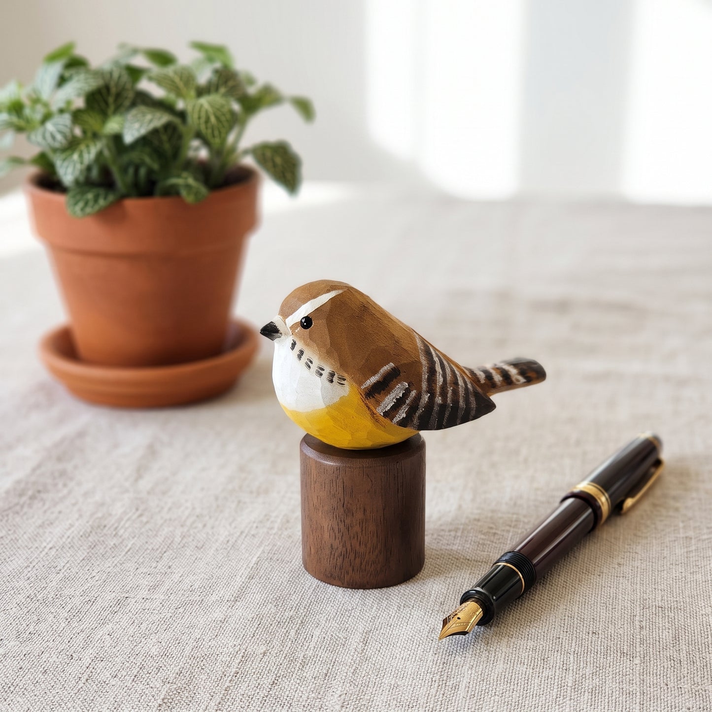 A hand-carved wooden Carolina Wren figurine perched on a small wooden block, styled on a linen surface next to a fountain pen, with a potted plant in the soft-focus background.
