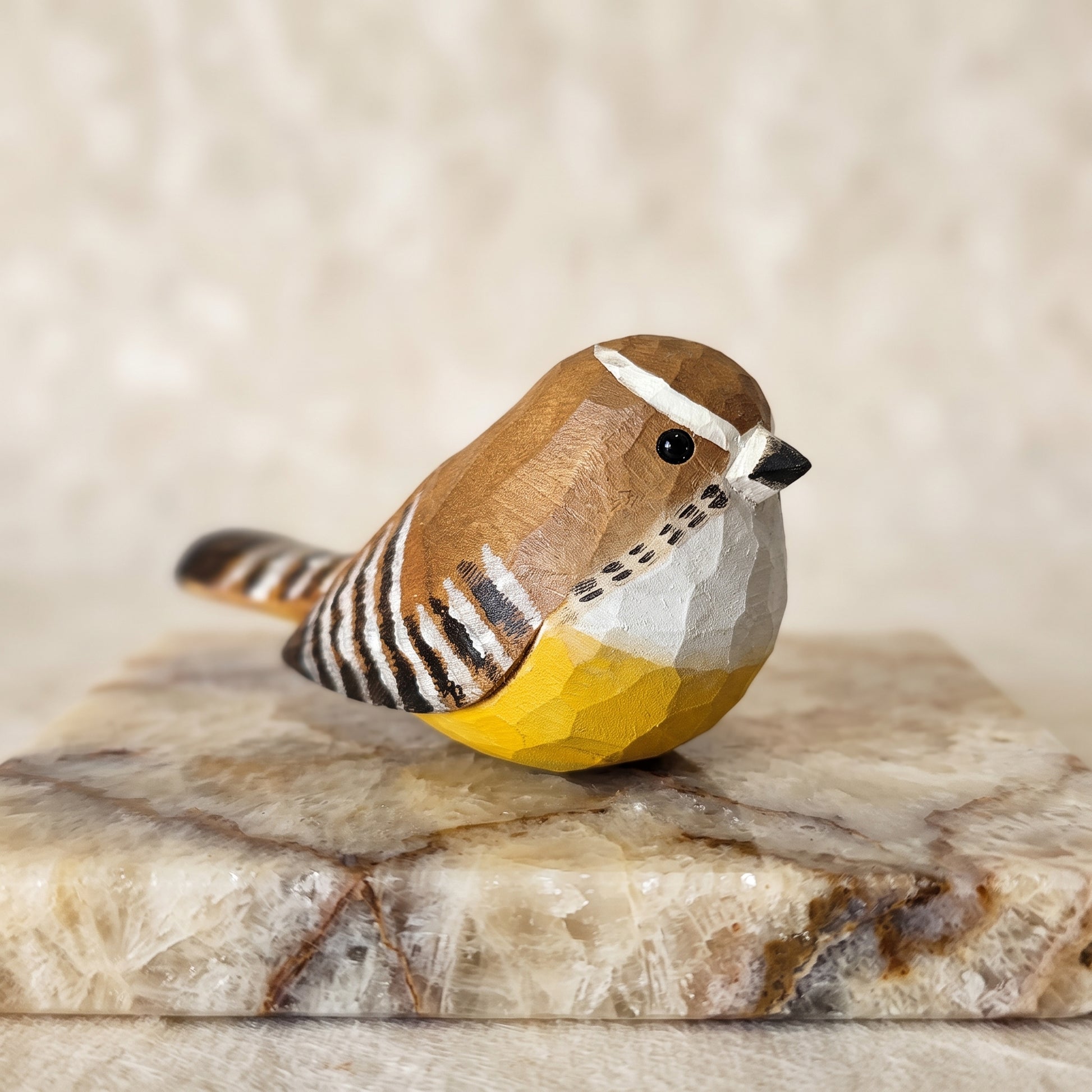 A small hand-carved wooden Carolina Wren figurine with a yellow belly, white and brown striped back, and black beak, displayed on a polished stone slab against a soft beige background.