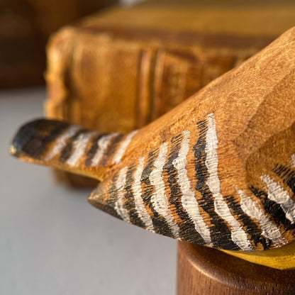 A close-up of a hand-carved wooden Carolina Wren figurine with painted black, white, and brown zebra-like stripes on its wing, resting on a wooden stand.