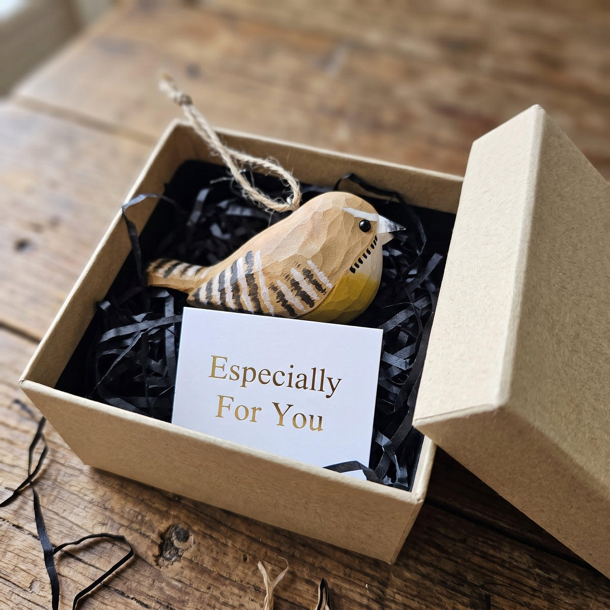 A wooden Carolina wren ornament nestled in black shredded paper inside a kraft gift box, with a white card reading 'Especially For You' in front. The box lid rests beside it on a rustic wooden table.