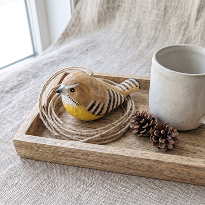 A carved wooden Carolina wren ornament with a yellow belly and black-and-cream striped wings, displayed on a rustic wooden tray with twine, pinecones, and a ceramic mug on a linen surface.