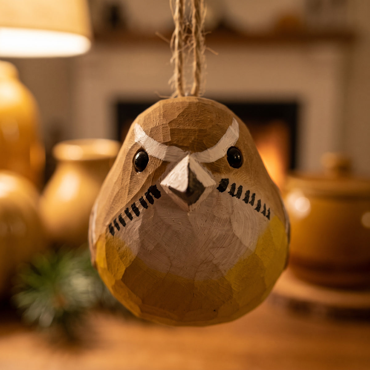 A handcrafted wooden Carolina wren ornament hanging by a twine string, featuring a rustic carved texture and painted details in yellow, brown, and white, set against a warm, softly blurred background with a fireplace and ceramic vessels.
