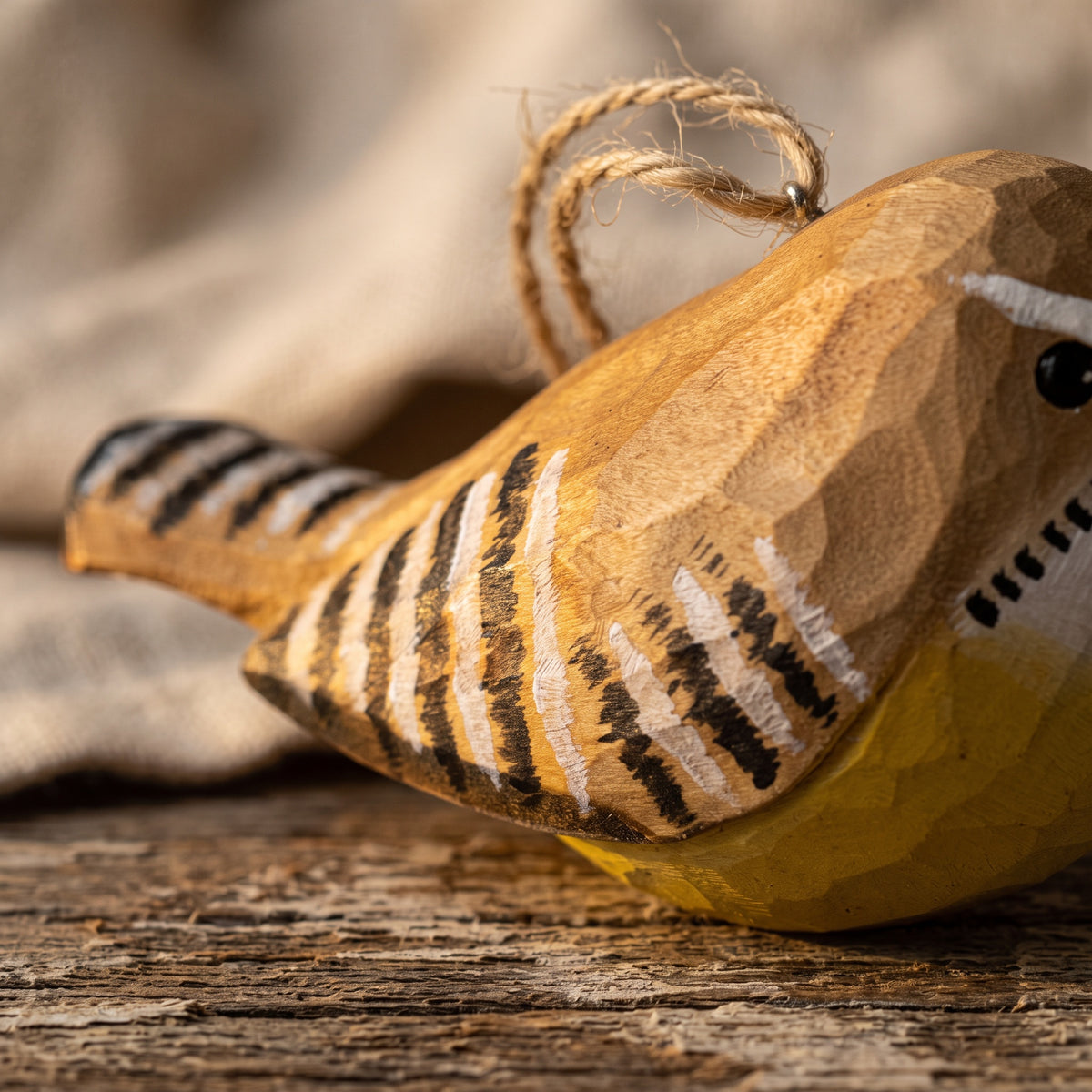 A close-up of a handcrafted wooden Carolina wren ornament with black and white painted stripes, a yellow belly, and a natural twine hanging loop, resting on a rustic wooden surface.