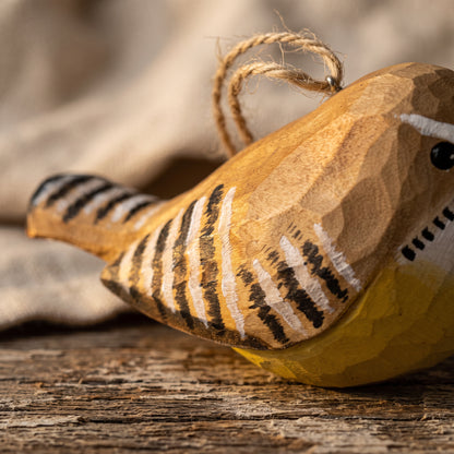 A close-up of a handcrafted wooden Carolina wren ornament with black and white painted stripes, a yellow belly, and a natural twine hanging loop, resting on a rustic wooden surface.