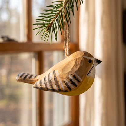 A handcrafted wooden Carolina wren ornament, painted in natural wood tones with black and white striped wing patterns, hanging from a pine branch in front of a softly blurred window.