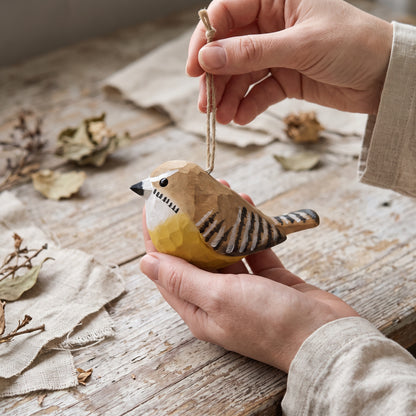 A handcrafted wooden Carolina wren ornament being held by two hands over a rustic wooden table with linen cloths and dried leaves. The ornament features a natural wood body with hand-painted yellow, white, and black details, and is suspended by a twine string.