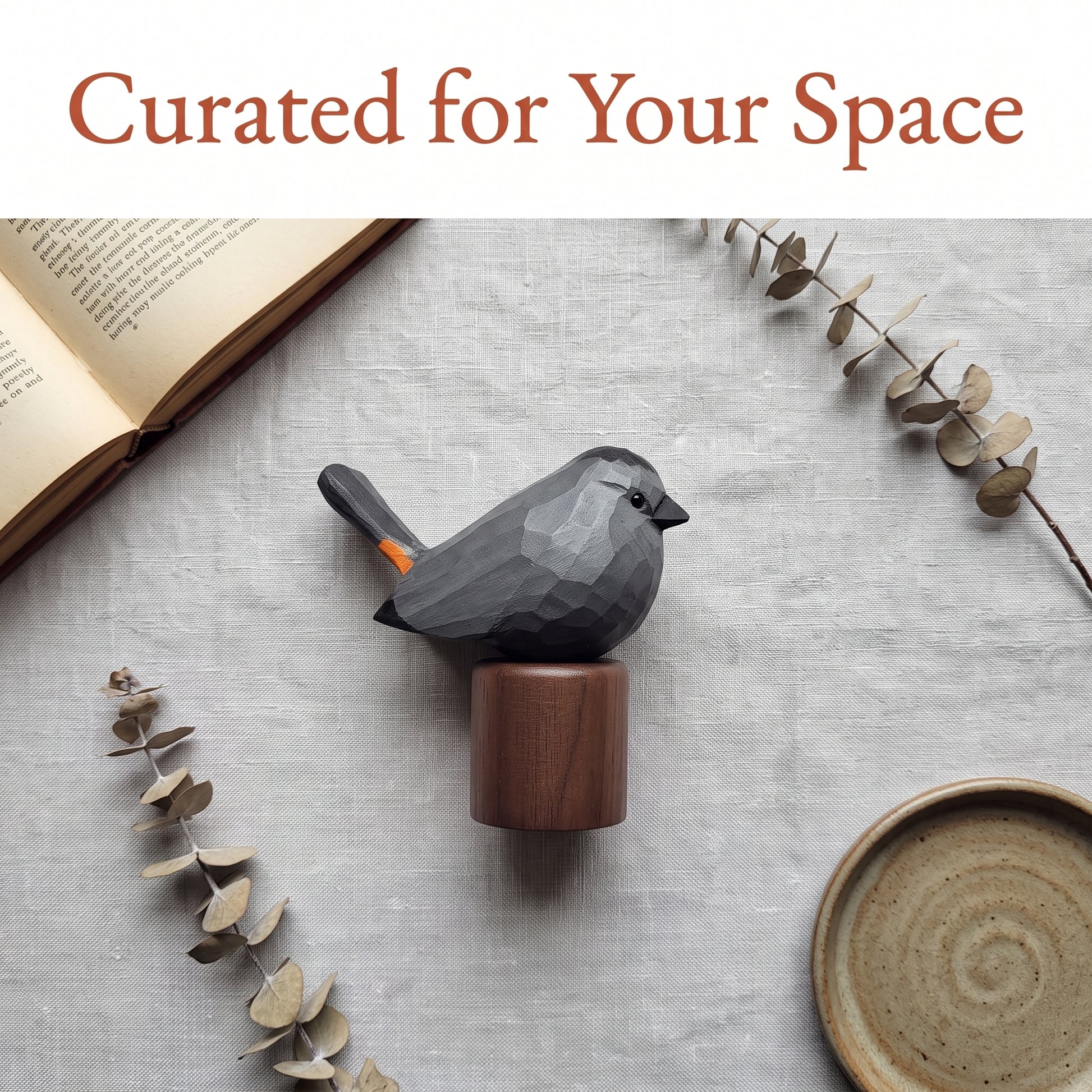 A faceted dark Gray Catbird figurine with a small orange accent on its tail, mounted on a round wooden base, styled on a light gray textured surface with dried eucalyptus stems, an open book, and a ceramic dish. The text 'Curated for Your Space' appears at the top.