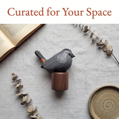A faceted dark Gray Catbird figurine with a small orange accent on its tail, mounted on a round wooden base, styled on a light gray textured surface with dried eucalyptus stems, an open book, and a ceramic dish. The text 'Curated for Your Space' appears at the top.