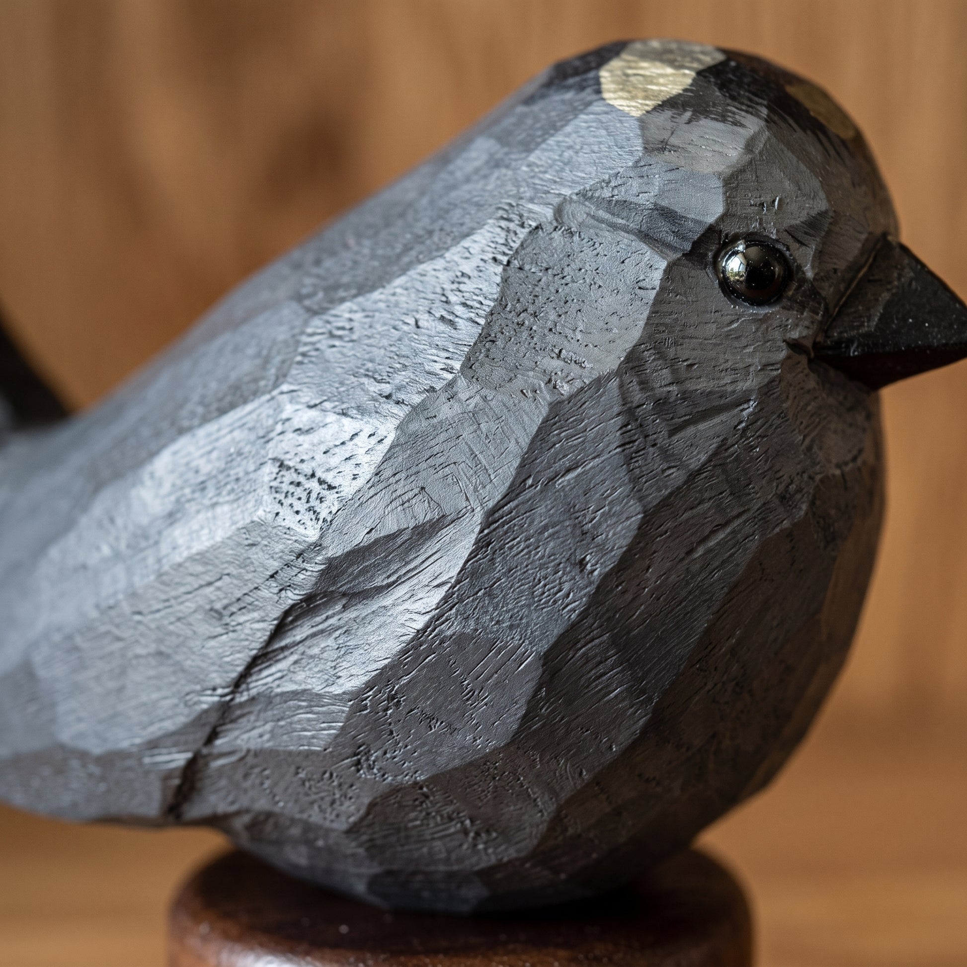 A close-up of a carved Gray Catbird sculpture with a faceted geometric design, finished in dark charcoal gray with a small gold accent on its head, perched on a round wooden base against a warm, blurred wood background.