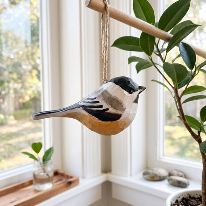 Hand-carved and hand-painted wooden Black-capped Chickadee ornament hanging from a jute string near a sunny window with house plants.