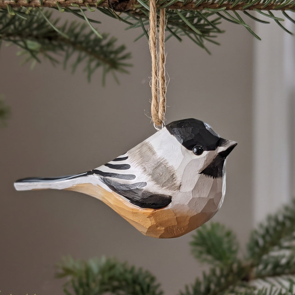 A handcrafted wooden bird ornament, shaped like a chickadee with a black, white, gray, and brown painted finish, hanging from a natural jute string on a pine tree branch.