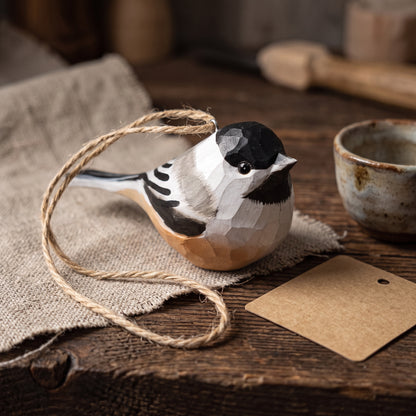 Hand-carved wooden Black-capped Chickadee ornament resting on burlap cloth next to a rustic cup, natural jute string, and a blank kraft tag.