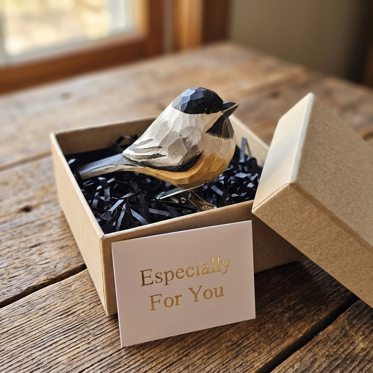 Hand-carved wood chickadee ornament displayed inside an open kraft gift box with black shredded paper and an "Especially For You" card on a rustic wood table.