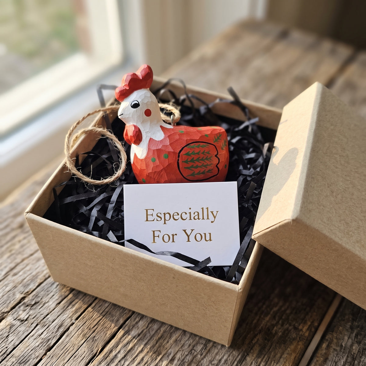 A small decorative rooster ornament nestled in black paper shred inside a kraft gift box, with a white card reading 'Especially For You' in front. The rooster is orange-red with a white face and red comb, and the box sits on a rustic wooden surface near a window.