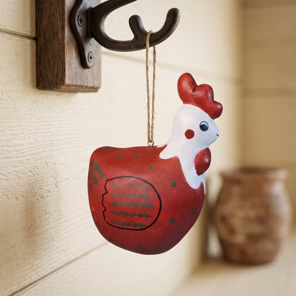 A hand-painted ceramic rooster ornament hanging from a rustic black metal hook on a wooden wall plaque. The rooster is primarily red with a white head and small green accents, set against a warm, softly blurred background.