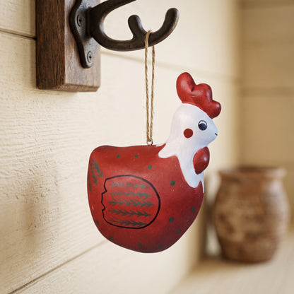 A hand-painted ceramic rooster ornament hanging from a rustic black metal hook on a wooden wall plaque. The rooster is primarily red with a white head and small green accents, set against a warm, softly blurred background.