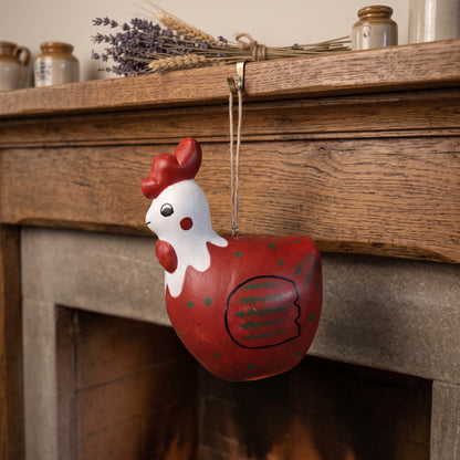 A hand-painted wooden hanging ornament shaped like a rooster chick, featuring a red body with green polka dots, a white head, and red comb and wattle, hanging from a string in front of a rustic wooden fireplace mantel.