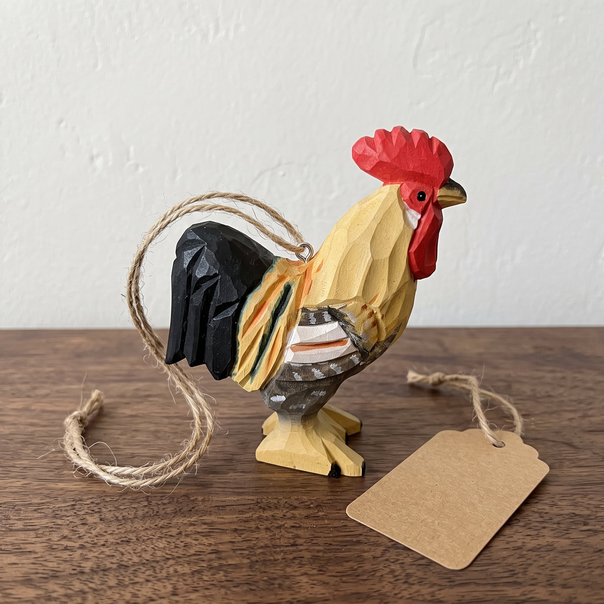 A hand-carved wooden rooster ornament with a red comb and wattle, yellow body, black tail feathers, and natural wood grain, hanging from twine with a blank kraft paper tag beside it on a wooden surface.