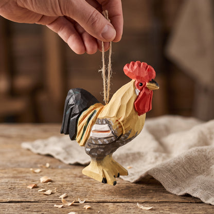 A hand holds a small hand-carved wooden rooster ornament by a twine loop, shown against a rustic wooden table with burlap fabric in the background.