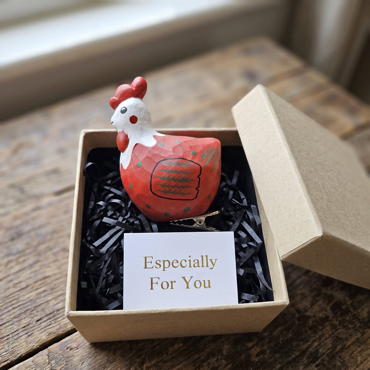 A hand-painted wooden rooster ornament in red, white, and black resting in an open kraft gift box filled with black shredded paper, featuring a small card reading "Especially For You" placed in the foreground.